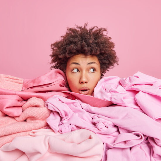 Shocked young Afro American woman shows only head buried in big heap of clothes looks away with stunned expression collects clothing for donation isolated over pink background. Wardrobe cleaning