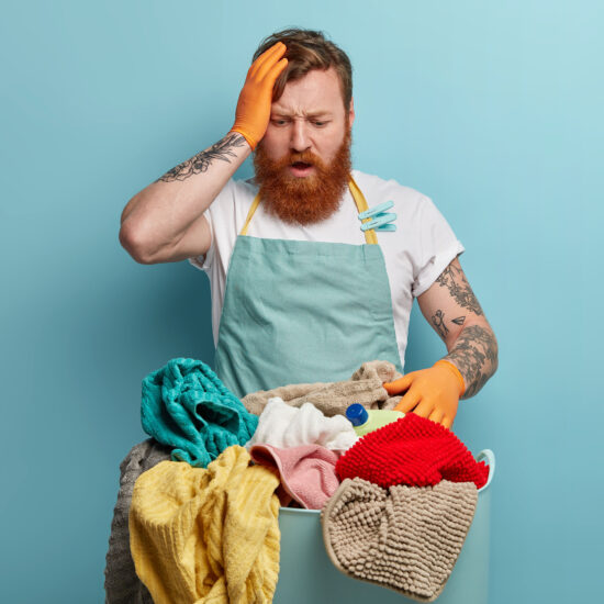 Frustrated overwhelmed puzzled foxy man has much work about house, keeps hand on head and stares at basket full of laundry, has washing time at home, doesnt know how to start, dressed in apron